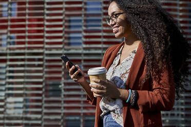 Smiling woman using smart phone holding reusable cup while standing by building - VEGF03121