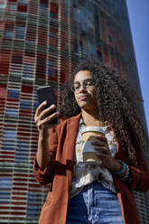 Woman using smart phone holding bamboo cup while standing against building in city - VEGF03123