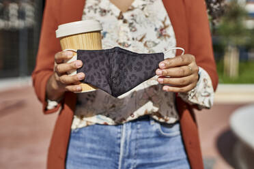 Woman holding bamboo mug and protective face mask standing in city on sunny day - VEGF03127