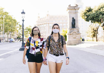 Young lesbian couple holding hands walking on street in city during COVID-19 - DGOF01592