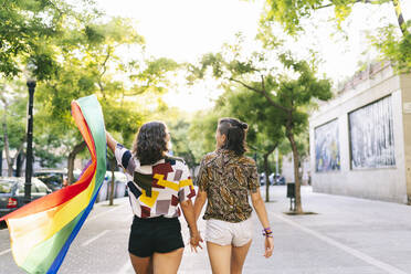Lesbisches Paar mit Regenbogenfahne hält sich auf dem Fußweg an den Händen - DGOF01621