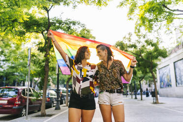 Lesbisches Paar, das sich gegenseitig anschaut, während es eine Regenbogenfahne auf einem Fußweg in der Stadt hält - DGOF01625