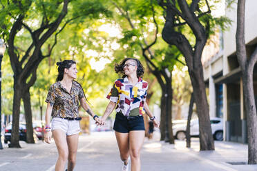 Glückliches lesbisches Paar hält sich an den Händen und läuft auf dem Fußweg in der Stadt an einem sonnigen Tag - DGOF01629
