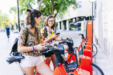 Freundinnen, die ein Mobiltelefon benutzen, während sie an einer Fahrradabstellanlage stehen - DGOF01632