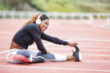 Female athlete listening music while doing stretching exercise sitting on runner track - GGGF00050