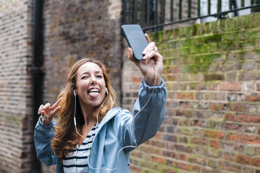 Carefree woman with in-ear headphones sticking tongue out while taking selfie through mobile phone in city - JMPF00527