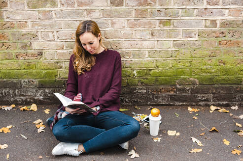 Woman reading book while sitting against brick wall on footpath - JMPF00530