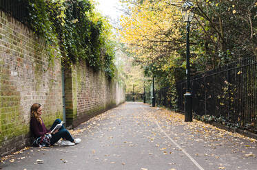 Woman reading book while sitting by brick wall on footpath during autumn - JMPF00531