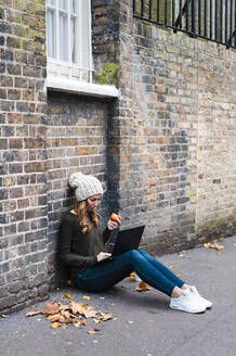 Frau, die einen Laptop benutzt, während sie auf dem Gehweg einen Apfel isst, der gegen eine Mauer gelehnt ist - JMPF00536
