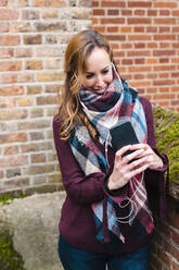 Woman with in-ear headphones using smart phone while standing against brick wall - JMPF00540