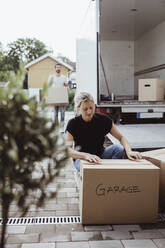Woman crouching by cardboard box while partner unloading luggage from moving van - MASF20431