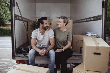 Lächelnde männliche und weibliche Partner sehen einander an, während sie an Pappkartons im Lieferwagen sitzen - MASF20449