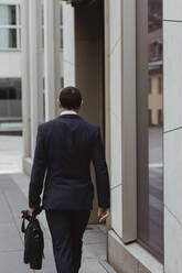 Rear view of businessman with bag walking by building in city - MASF20474