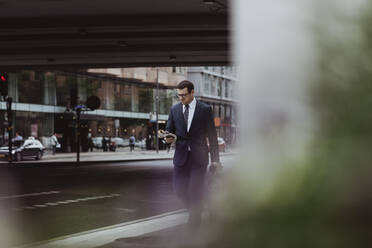 Businessman with bag using smart phone while walking in city - MASF20476