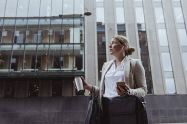 Female entrepreneur with bag and coffee cup looking away while standing against building in city - MASF20478
