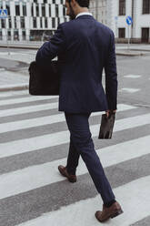 Rückansicht eines männlichen Unternehmers mit Tasche und Ordner beim Überqueren der Straße n Stadt - MASF20482