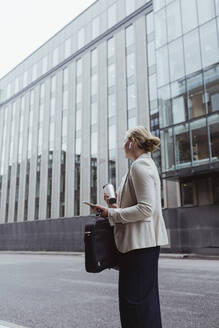 Seitenansicht eines Unternehmers mit Tasche und Kaffeetasse, der gegen ein Gebäude in der Stadt steht - MASF20490