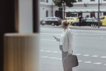 Seitenansicht eines Unternehmers mit Tasche und Mobiltelefon, der in der Stadt steht und dabei wegschaut - MASF20495