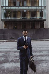 Männlicher Unternehmer mit Tasche, der ein Smartphone benutzt, während er in der Stadt steht - MASF20499