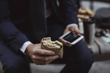 Mittelteil eines Unternehmers mit Smartphone und Sandwich in der Stadt sitzend - MASF20511