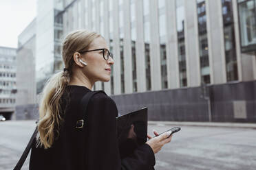 Rear view of businesswoman with in-ear headphones holding file in city - MASF20527