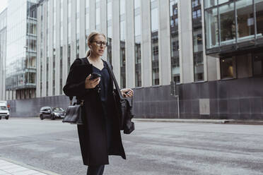 Contemplating businesswoman with smart phone walking in city - MASF20530