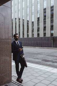 Unternehmer mit Tasche mit Smartphone in der Hand, während er in der Stadt steht - MASF20542