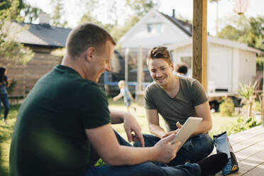 Seitenansicht eines lächelnden Mannes, der seinem homosexuellen Partner ein digitales Tablet zeigt, während Mütter und Sohn im Garten spielen - MASF20550