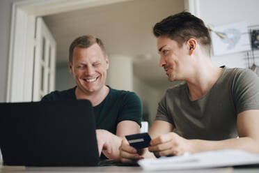 Smiling homosexual couple paying bill through laptop on dining table at home - MASF20551