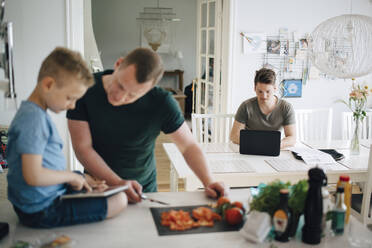 Mann benutzt Laptop am Esstisch, während Vater und Sohn das digitale Tablett auf der Küchentheke betrachten - MASF20554