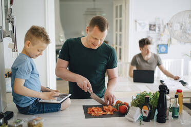 Father cutting tomato while son using digital tablet on kitchen counter - MASF20555