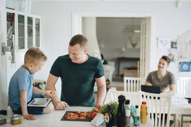 Smiling father talking to son holding tomato while using digital tablet on kitchen counter - MASF20557