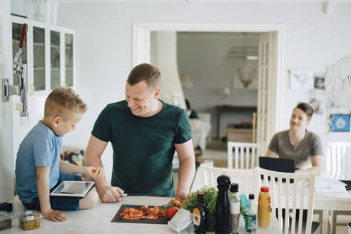 Lächelnder Vater spricht mit Sohn, der eine Tomate in der Hand hält, während er ein digitales Tablett auf der Küchentheke benutzt - MASF20557