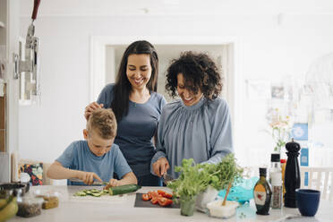 Smiling women looking at son cutting cucumber in kitchen - MASF20561