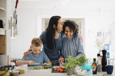 Smiling woman kissing partner while son cutting cucumber in kitchen - MASF20562