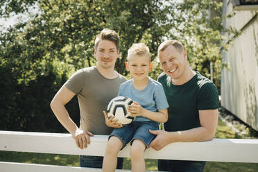 Portrait of homosexual fathers with son holding soccer ball in yard - MASF20583