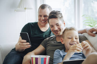 Lächelnde Väter und Söhne, die die Technik nutzen, während sie zu Hause auf dem Sofa Popcorn essen - MASF20593