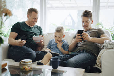 Lächelnder Vater mit Smartphone, der seinen Sohn mit einem digitalen Tablet auf dem Sofa zu Hause anschaut - MASF20595