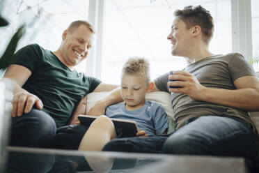 Lächelnde homosexuelle Väter sprechen, während der Sohn zu Hause ein digitales Tablet benutzt - MASF20596