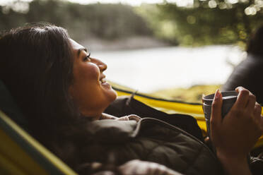 Lächelnde Frau mit Kaffee, die im Urlaub auf einer Hängematte im Wald wegschaut - MASF20640