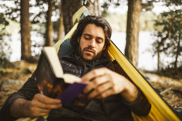 Junger Mann genießt das Lesen von Büchern, während er auf einer Hängematte im Wald liegt - MASF20651