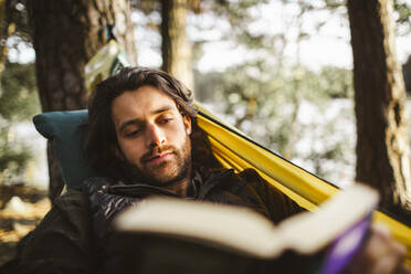 Young man reading book while lying on hammock in forest - MASF20653