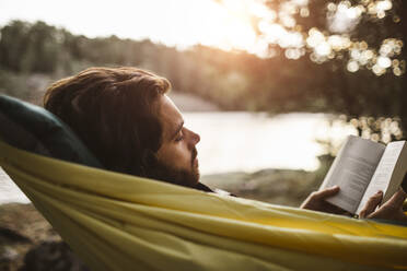 Betrachtender Mann liest Buch, während er über einer Hängematte im Wald liegt - MASF20656