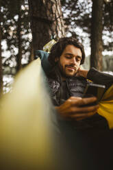 Junger Mann benutzt Mobiltelefon, während er über einer Hängematte im Wald liegt - MASF20657