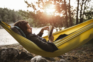 Contemplating man reading book while lying down over hammock in forest - MASF20659