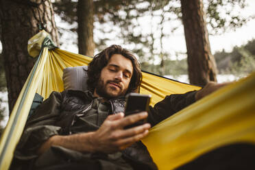 Junger Mann benutzt Smartphone, während er über einer Hängematte im Wald liegt - MASF20660