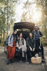 Porträt von lächelnden Freundinnen und Freunden, die während des Urlaubs im Kofferraum eines Autos sitzen - MASF20679