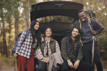 Lächelnde männliche und weibliche Freunde sitzen während des Urlaubs im Kofferraum - MASF20680