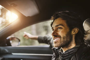 Lächelnder Mann mit der Hand vor dem Autofenster, der während einer Autofahrt wegschaut - MASF20693