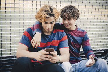 Lächelnde Freunde benutzen Smartphone, während sie in der Stadt gegen das Netz sitzen - MASF20768